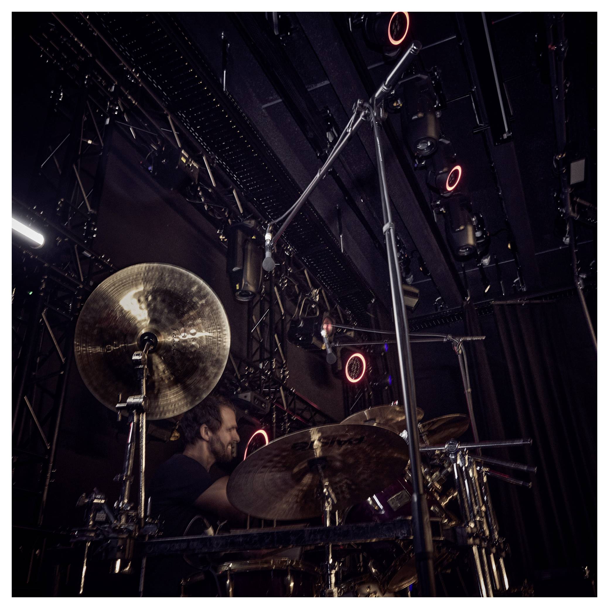 Un batteur joue sur une scène faiblement éclairée, entouré de cymbales et de microphones, avec des projecteurs de scène et une structure en arrière-plan.