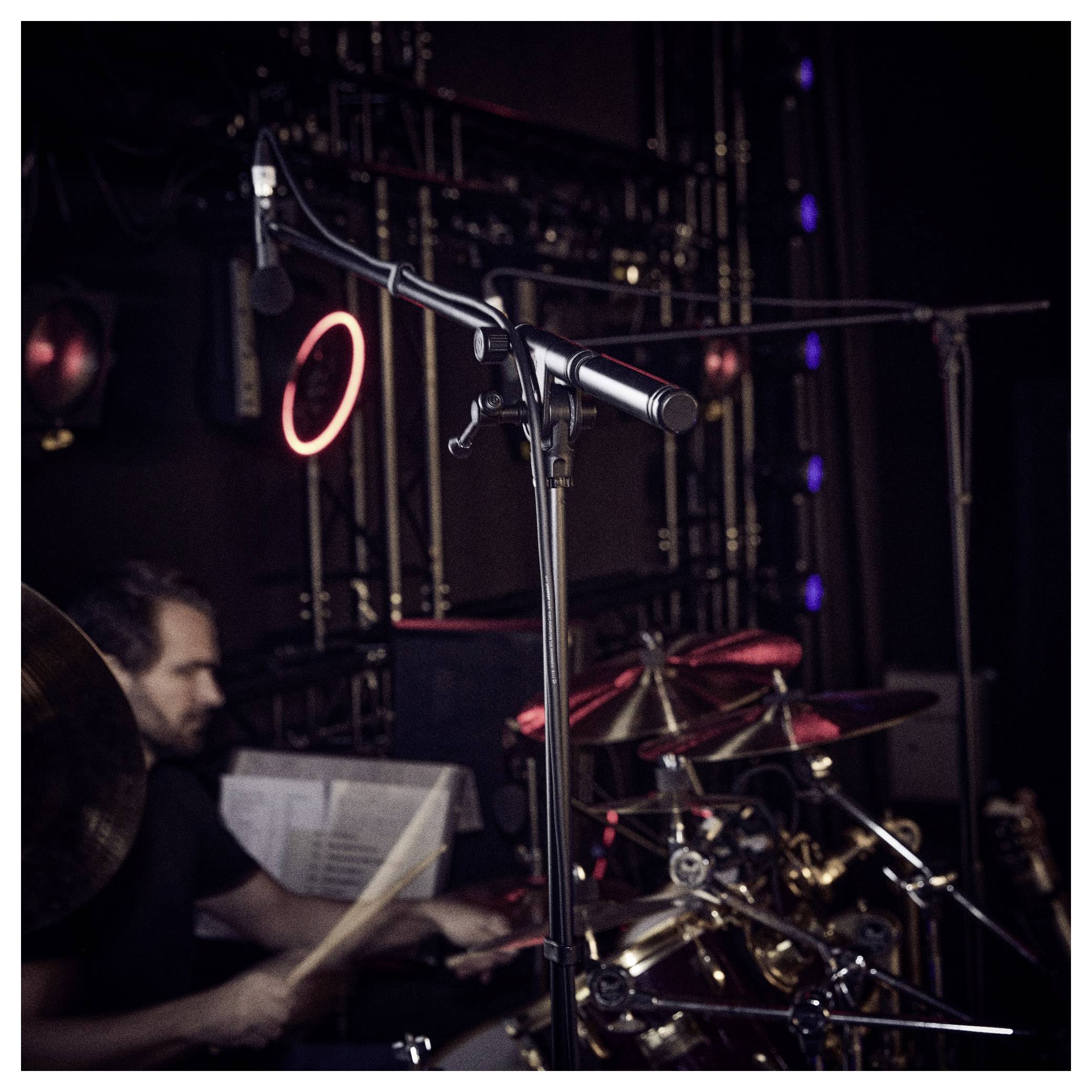 Un batteur joue sur une batterie sur scène. Un microphone est positionné au-dessus, capturant la performance. L'ambiance est un lieu de concert faiblement éclairé.