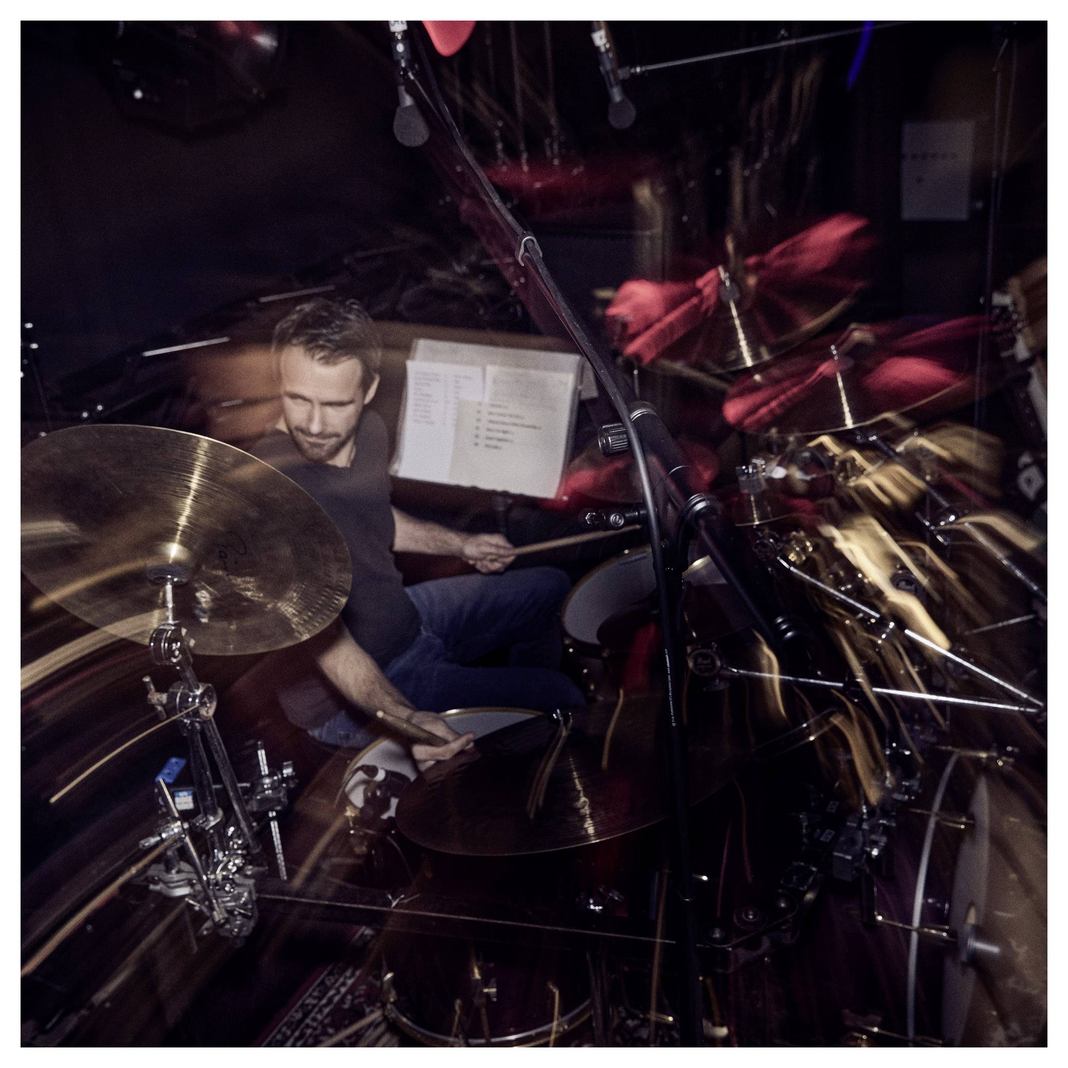 Un batteur joue énergiquement d'une batterie dans un studio d'enregistrement faiblement éclairé, entouré de microphones et d'équipements sonores.