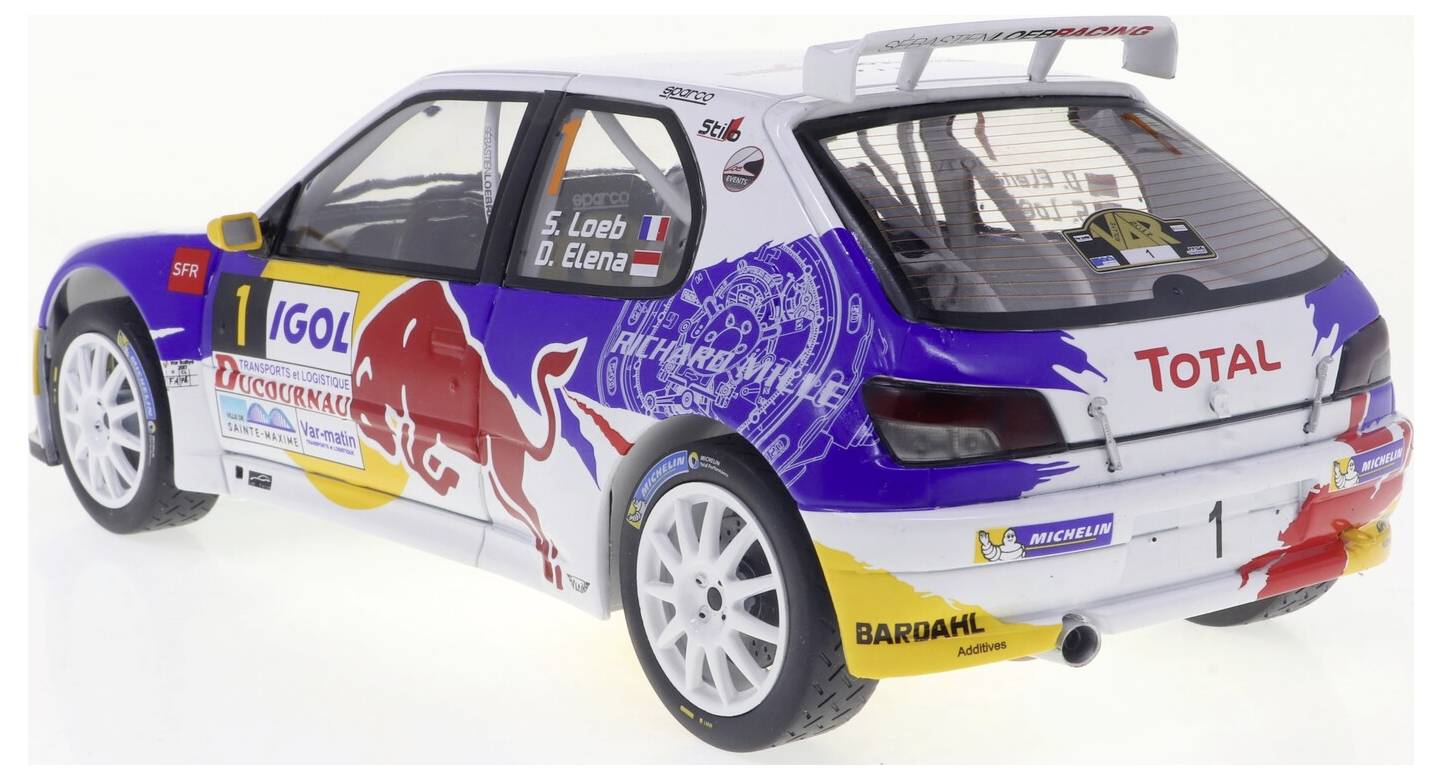 Une voiture de rallye avec des graphismes Rouge Bull vibrants, arborant un aileron et des logos incluant 'Total' et 'Michelin', est montrée depuis l'arrière côté.