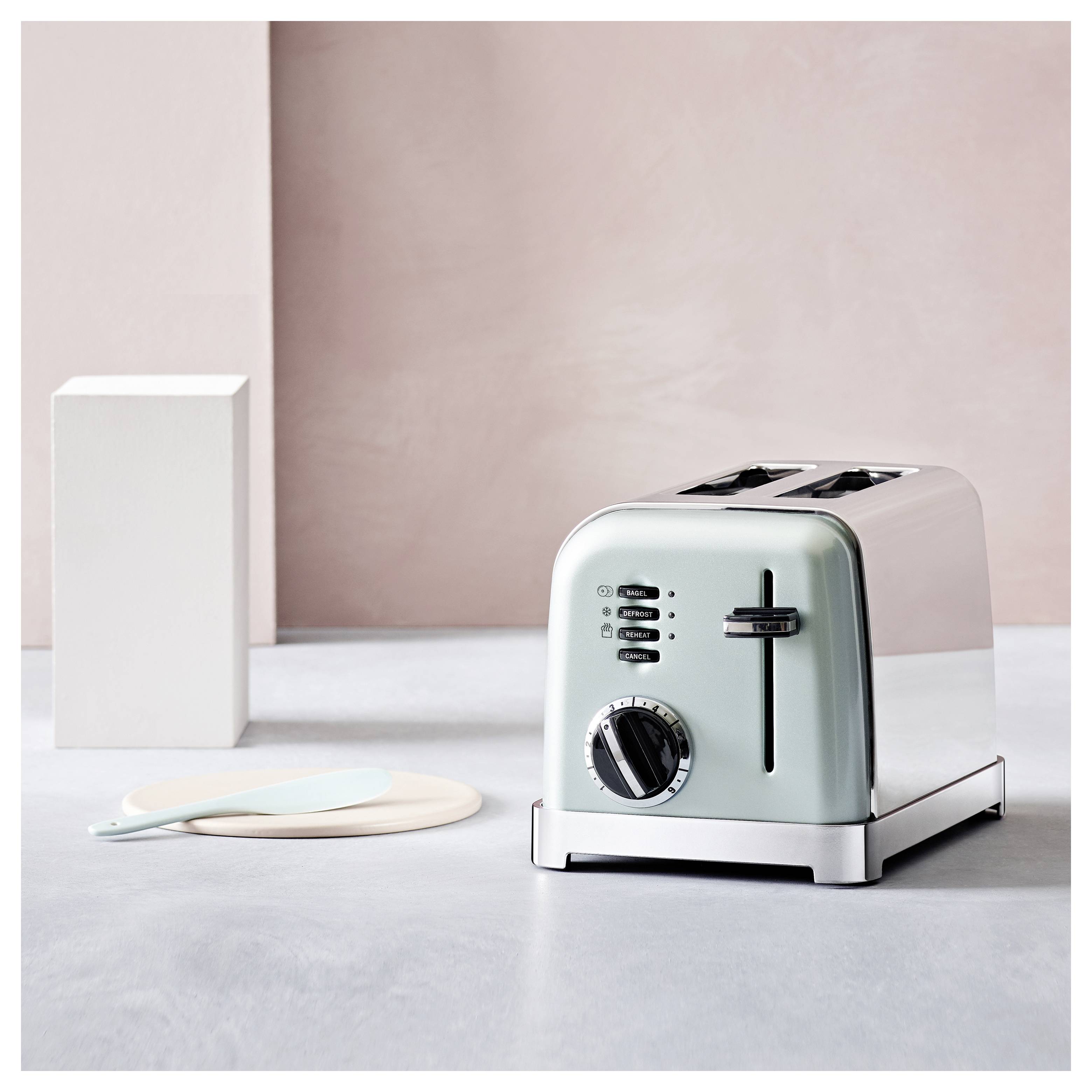 Un grille-pain élégant, argenté à deux fentes, posé sur un comptoir avec un design minimaliste, à côté d'un étendeur de couleur vert pastel et un bloc blanc en arrière-plan.