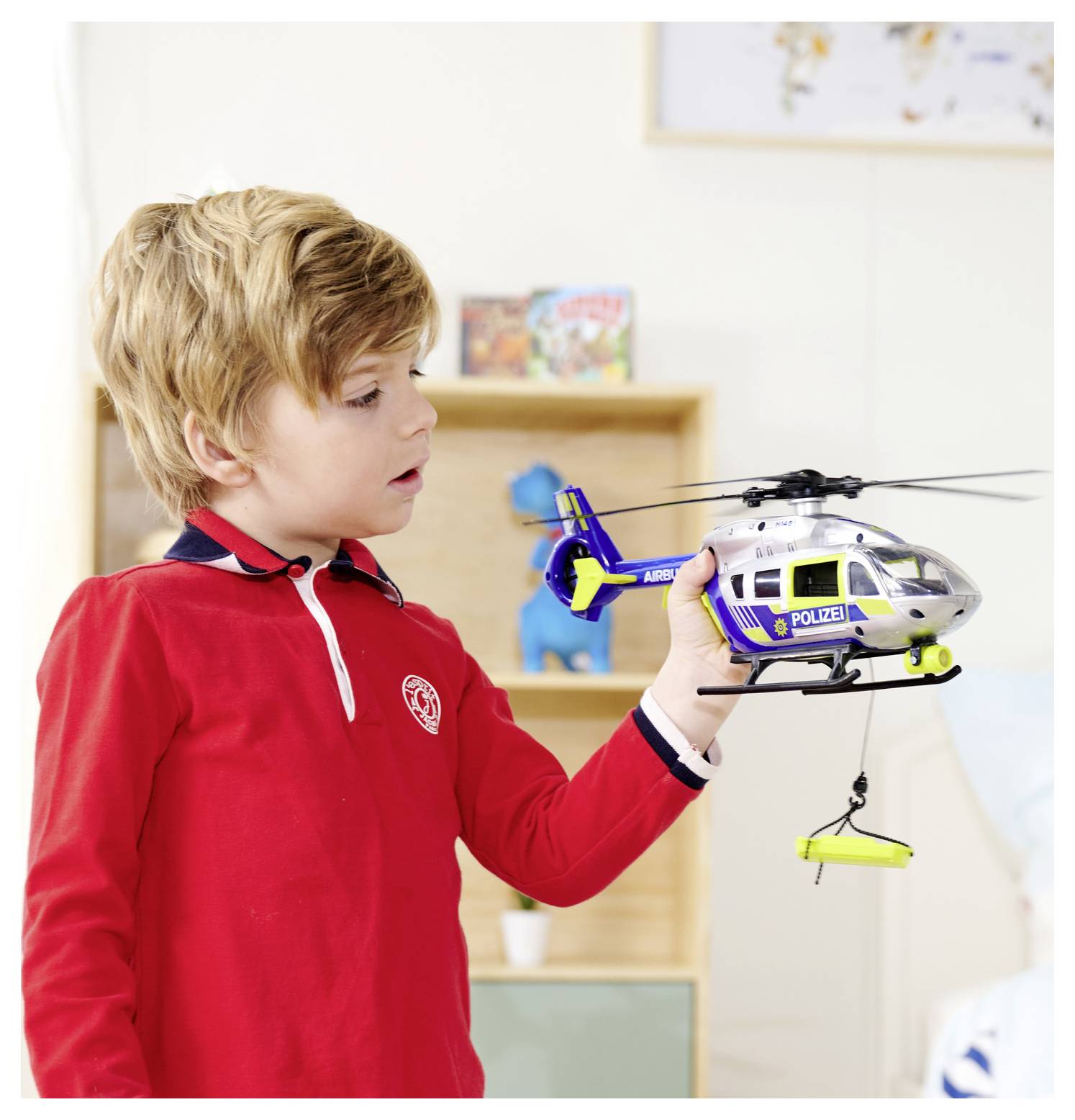 Un enfant portant un t-shirt rouge examine un hélicoptère de police miniature dans une pièce avec des étagères et des jouets en arrière-plan.