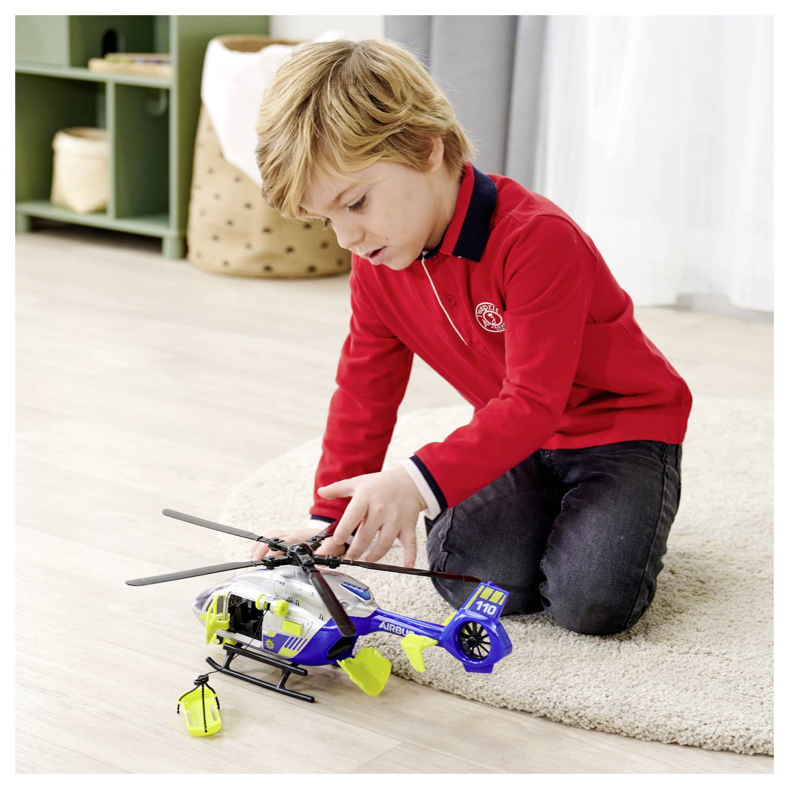 Un jeune garçon portant un pull rouge joue avec un hélicoptère jouet bleu et blanc sur un tapis beige dans un décor de chambre douillet.