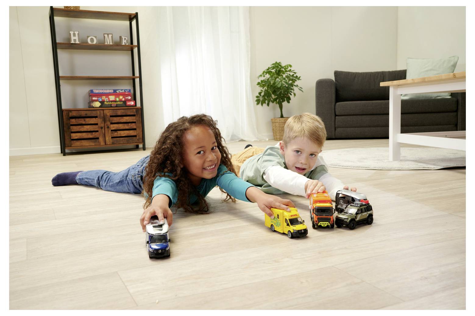 Deux enfants, une fille et un garçon, sont allongés sur le sol dans un salon, poussant des voitures miniatures de manière ludique. Un canapé et une étagère sont visibles en arrière-plan.
