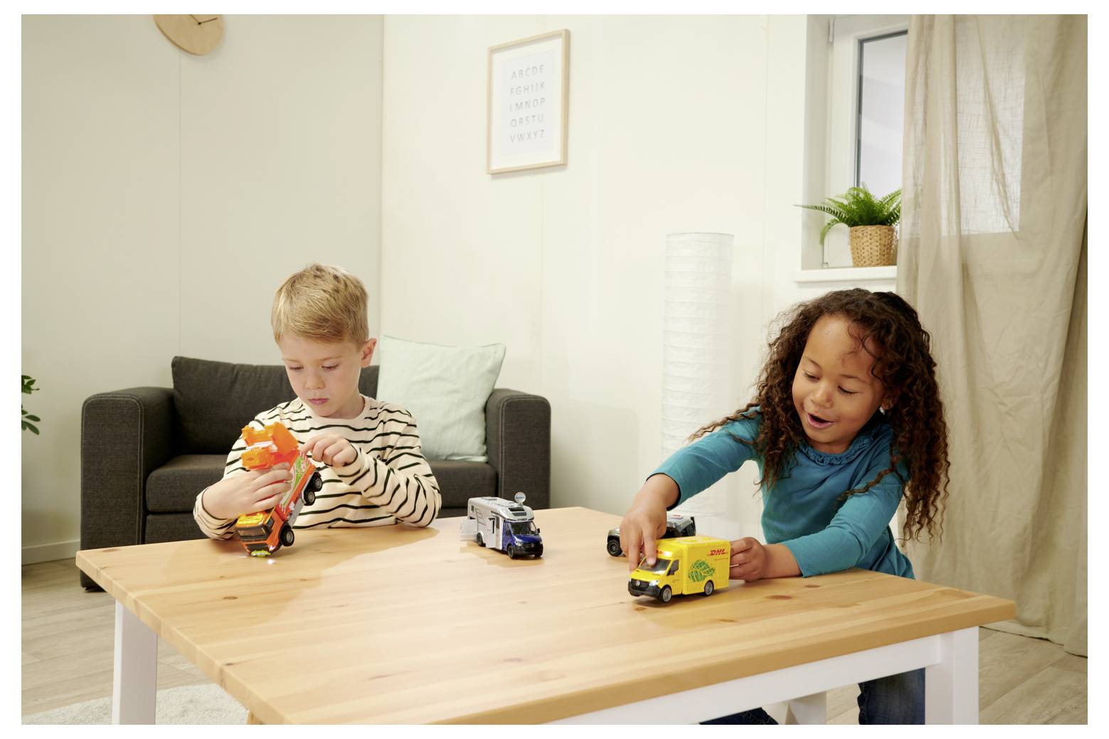 Un garçon et une fille jouent avec des camions miniatures sur une table en bois dans un salon douillet, avec un canapé gris et des murs de couleur claire.