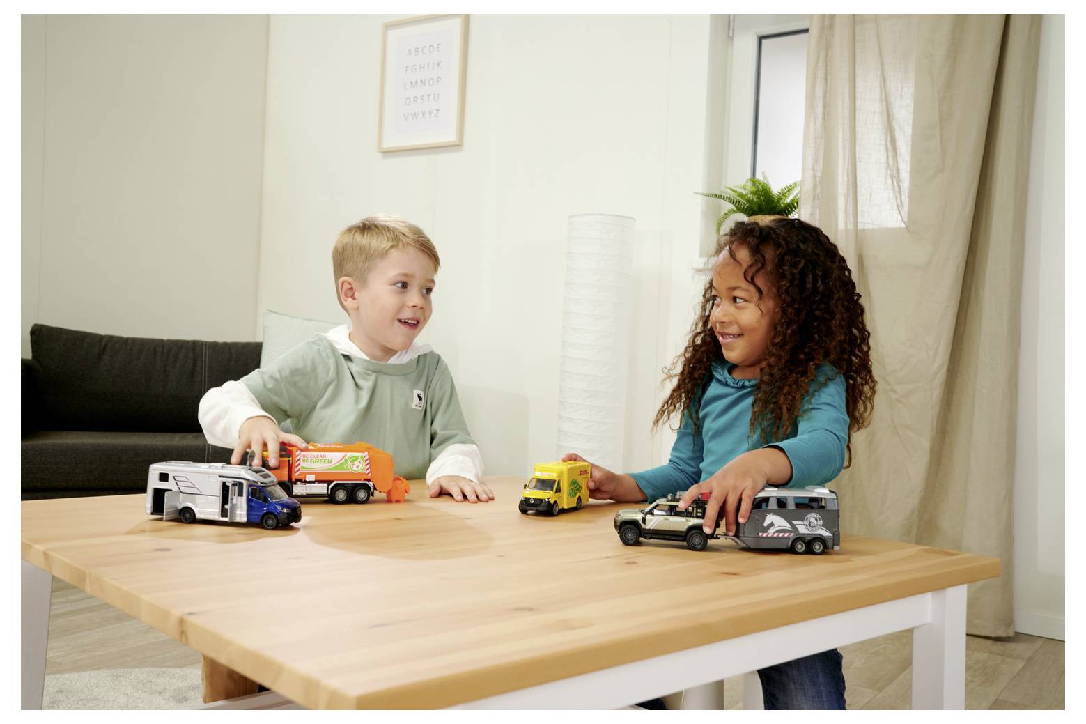 Deux enfants sourient en jouant avec des camions jouets sur une table en bois dans un décor de salon.