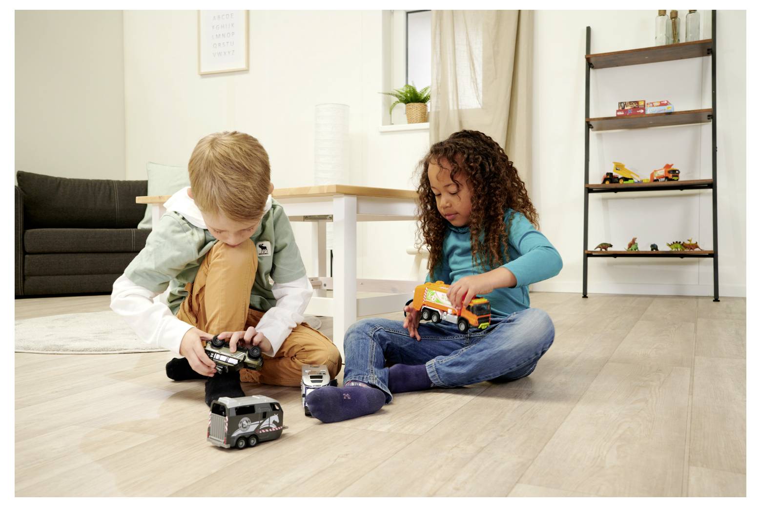 Deux enfants, un garçon et une fille, sont assis sur le sol en bois et jouent avec des camions miniatures dans un salon avec un canapé et une étagère en arrière-plan.