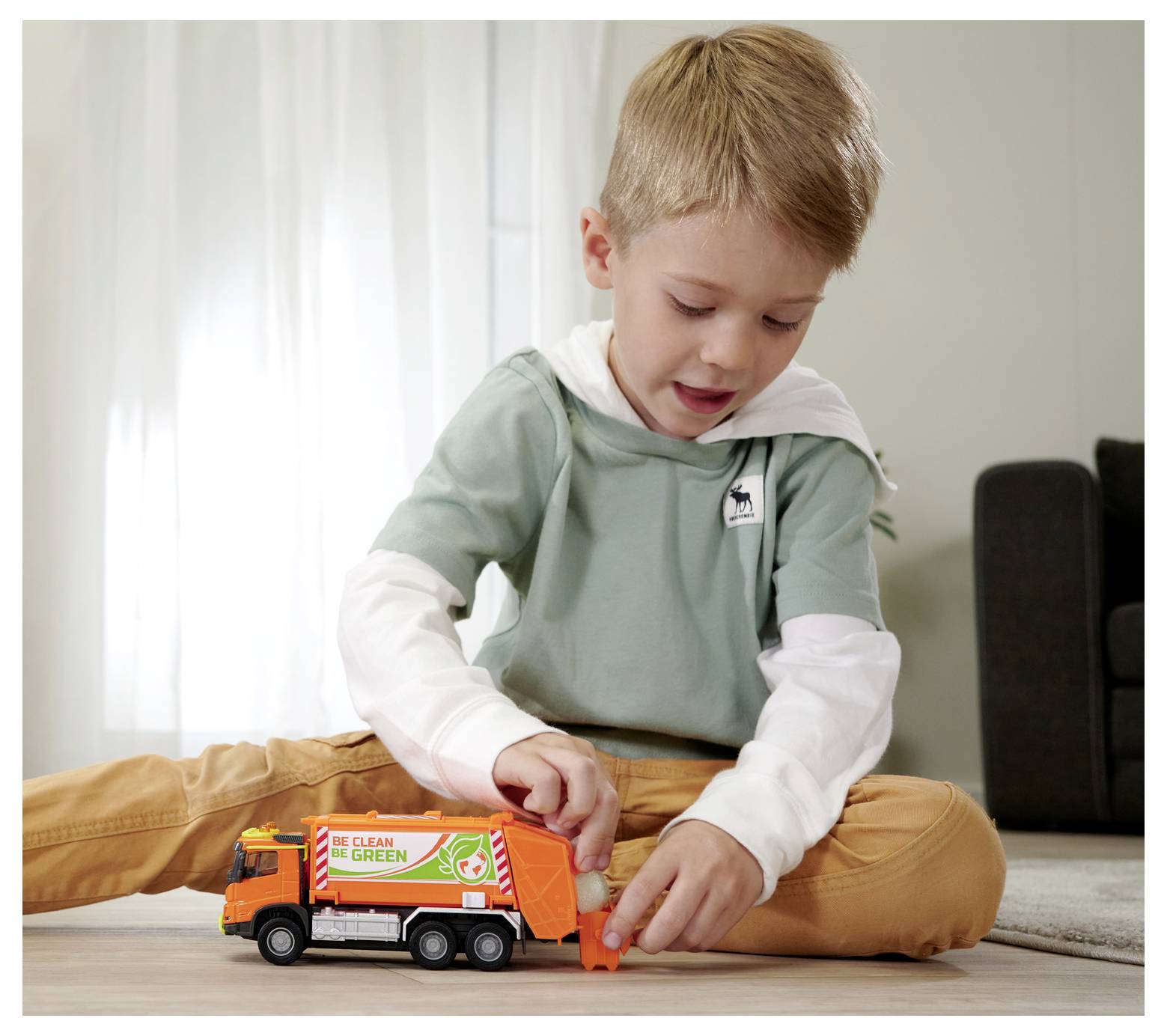 Un garçon est assis par terre, jouant avec un camion poubelle orange sur lequel est inscrit « Clean Green ». La pièce est doucement éclairée, avec une chaise en arrière-plan.