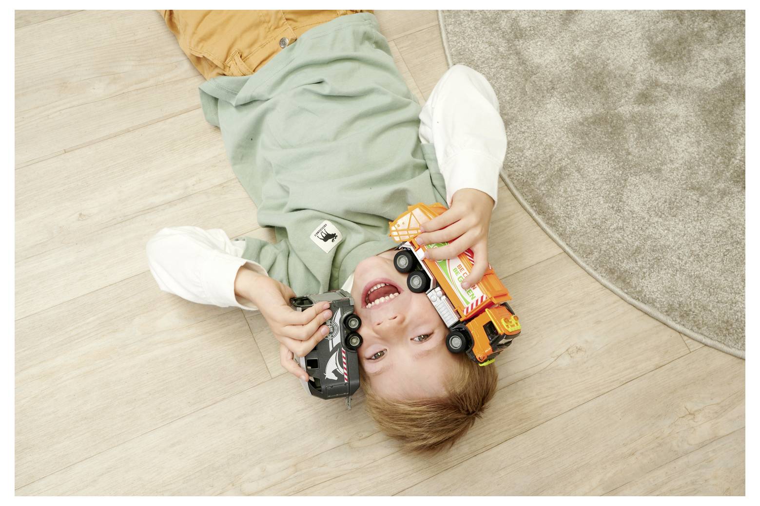 Un enfant allongé sur un plancher en bois, souriant, tenant deux camions jouets, l'un orange et l'autre noir, près d'un tapis circulaire gris.