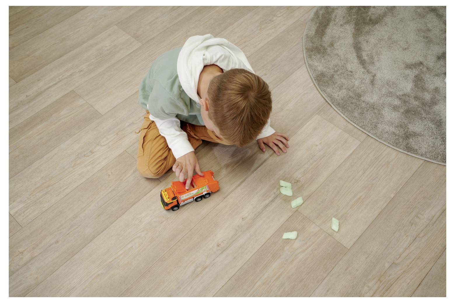 Un enfant joue sur le sol en bois avec un camion jouet orange et des blocs de mousse éparpillés à proximité. Un tapis rond gris est partiellement visible.