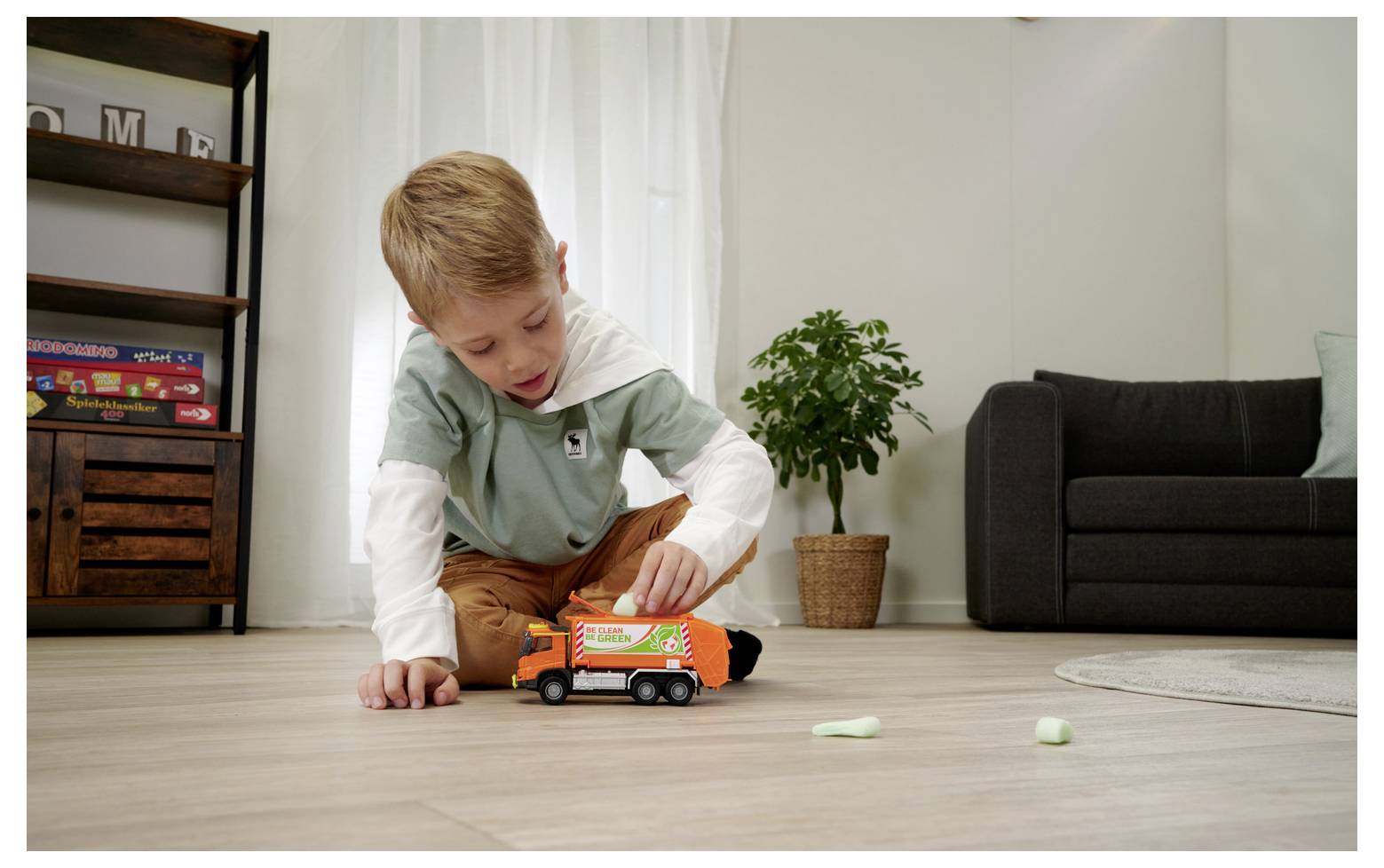 Un jeune enfant joue sur le sol avec un camion poubelle miniature dans un décor de salon, avec un canapé, une plante et une bibliothèque.
