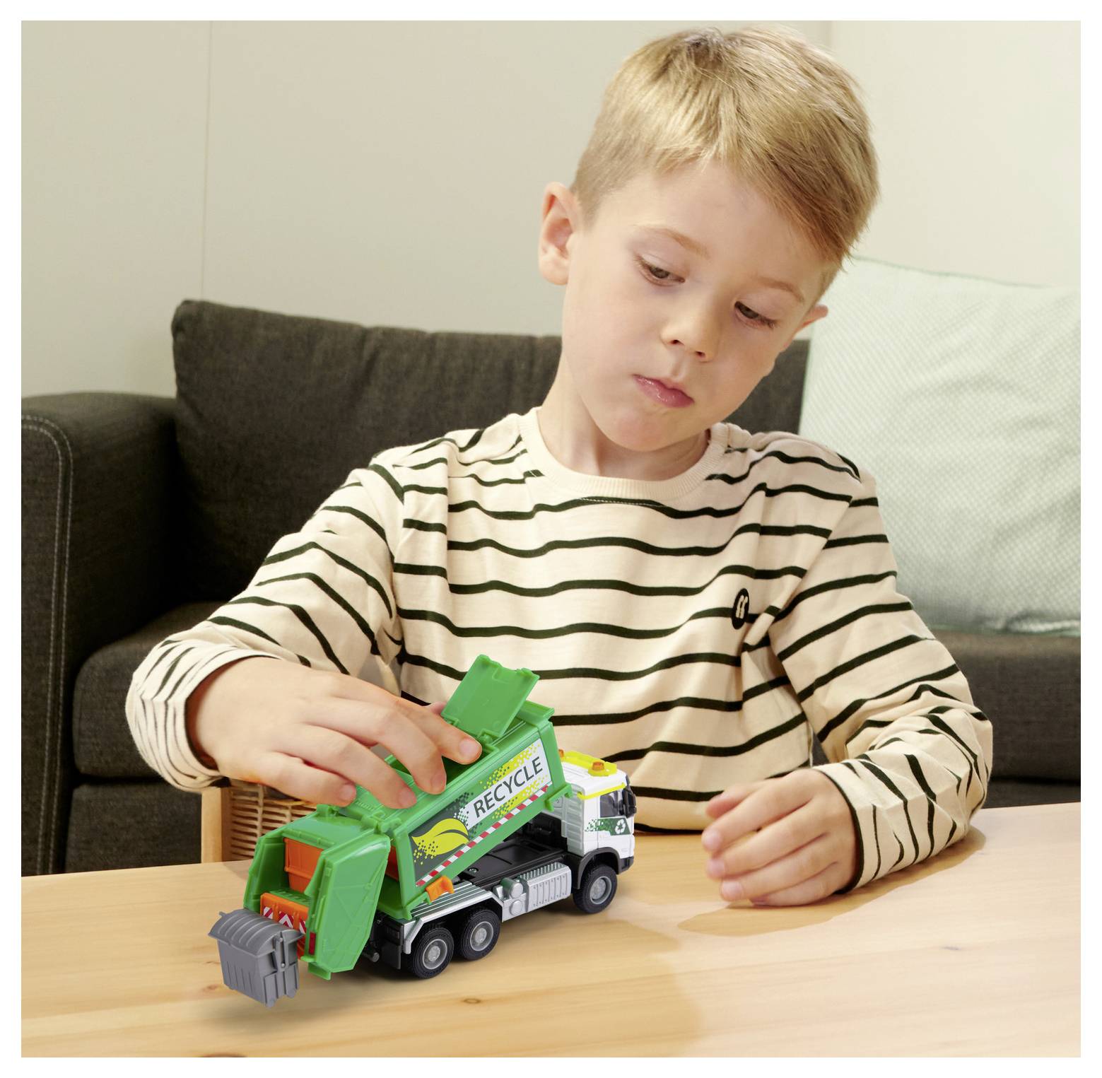 Un jeune garçon portant un t-shirt rayé joue avec un camion-poubelle miniature sur une table en bois, s'adonnant à un jeu imaginaire dans un décor de salon.