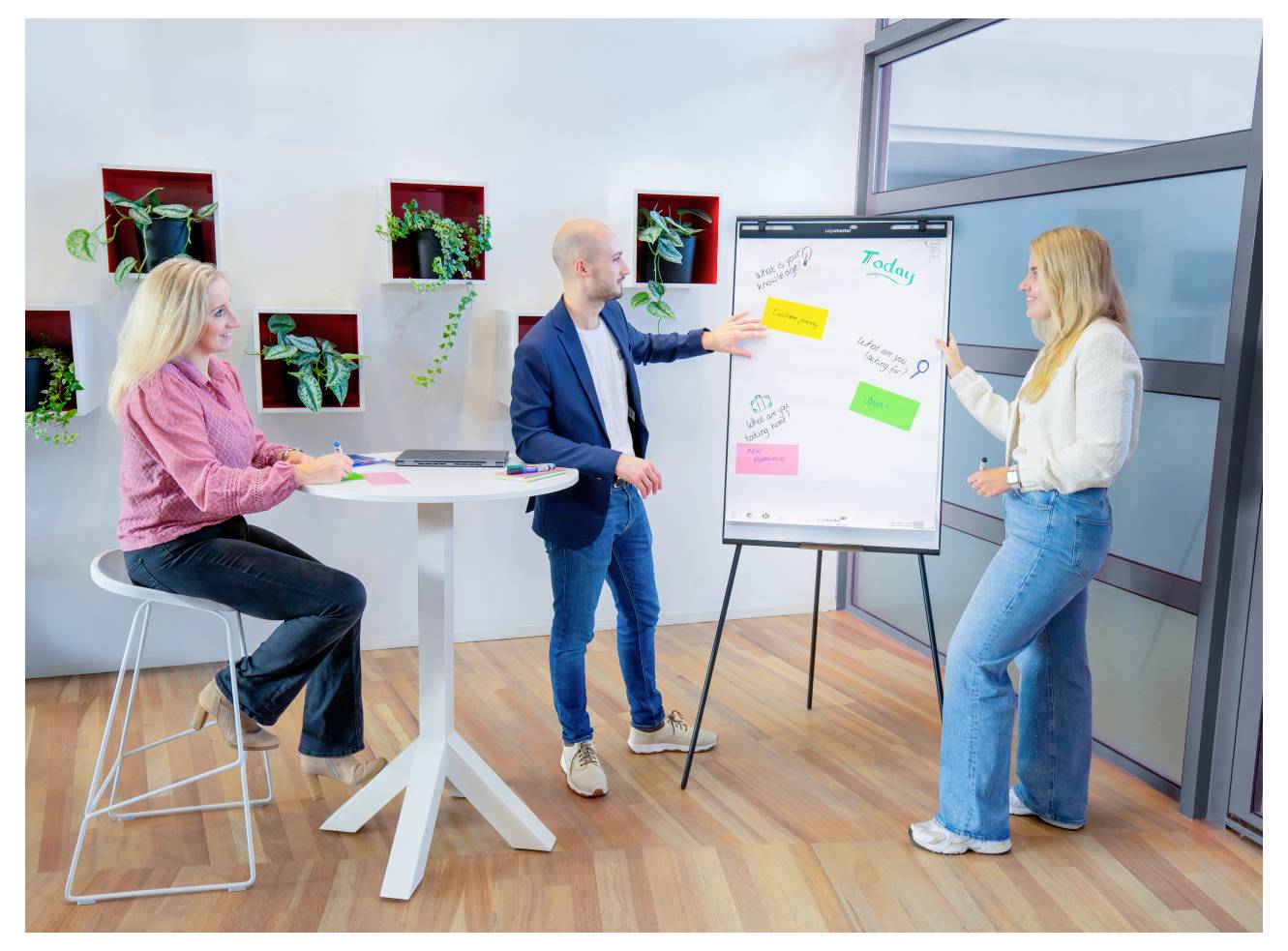 Trois personnes discutent des idées dans un bureau. L'un d'entre eux pointe du doigt un tableau blanc couvert de notes autocollantes. Les autres écoutent attentivement.