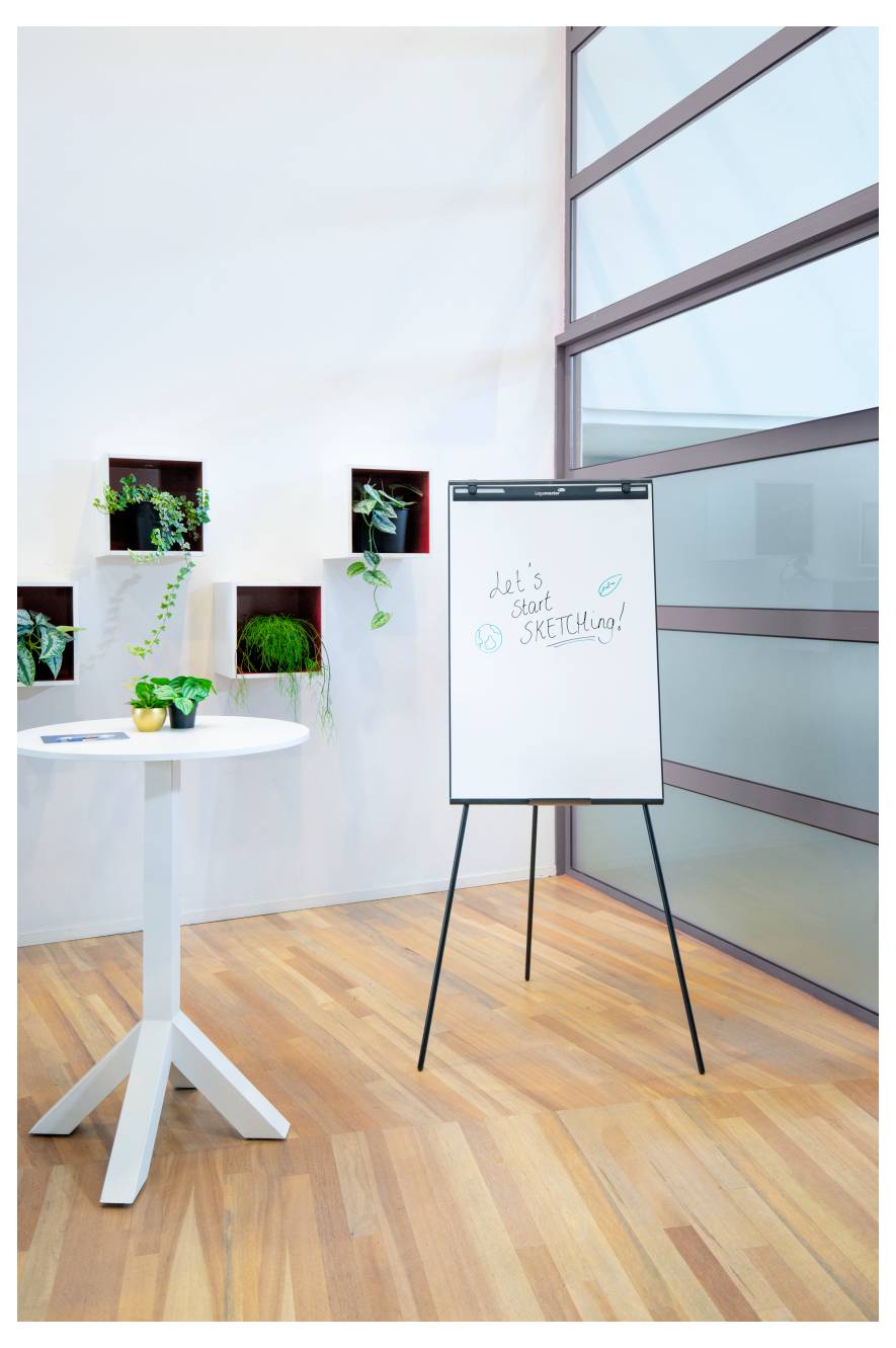 Un coin de bureau moderne avec un tableau à feuilles mobiles sur pied portant l'inscription « Commençons à DESSINER ! », une table ronde avec une plante, et de la verdure murale.