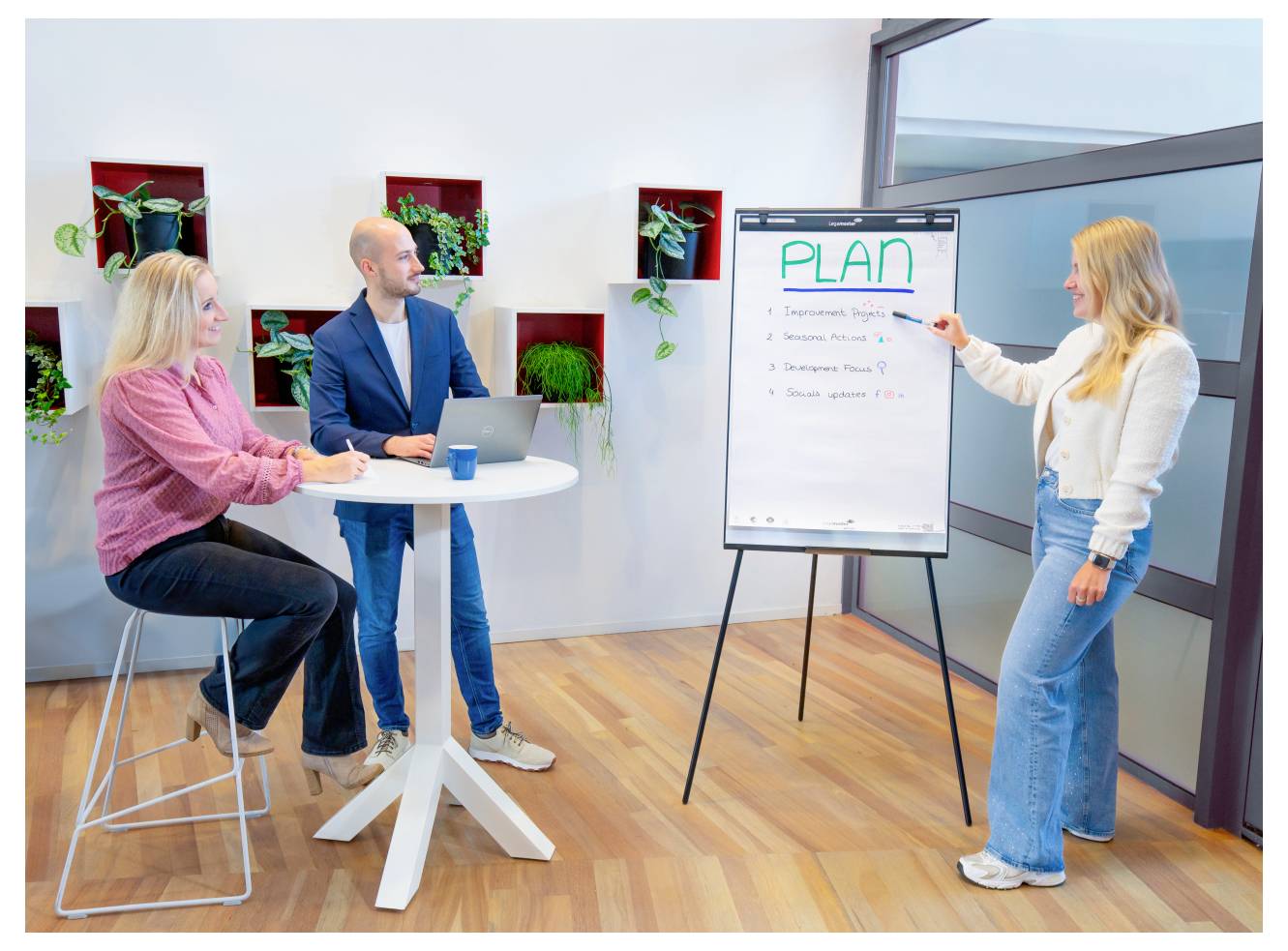 Une femme présente un tableau à feuilles mobiles intitulé 'PLAN' avec des points clés à deux collègues assis dans un bureau moderne avec des plantes murales, discutant de stratégies.