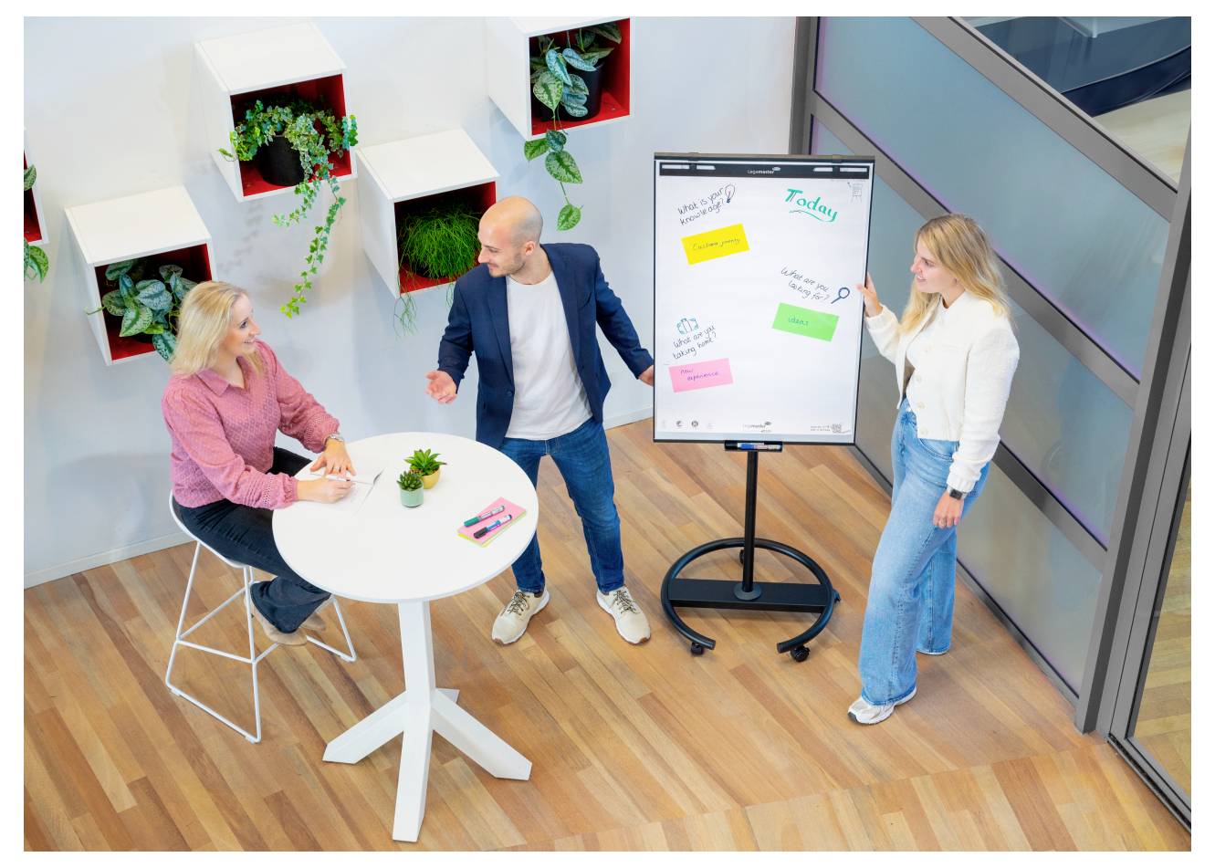 Trois personnes discutent des idées dans un espace de bureau, avec deux personnes debout à côté d'un tableau à feuilles et une personne assise à une table ronde.