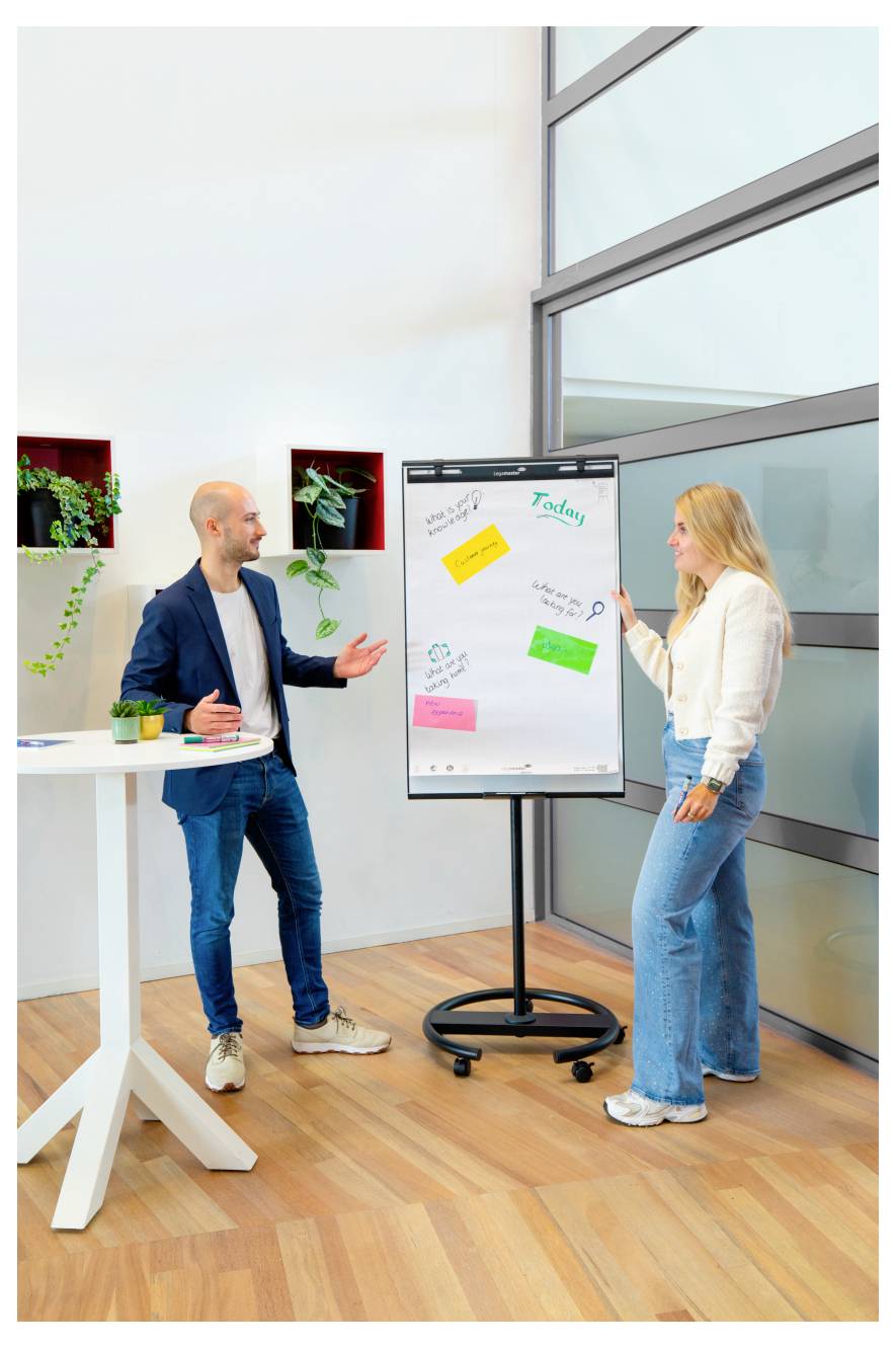 Un homme et une femme discutent d'idées dans un bureau moderne, pointant des notes autocollantes colorées sur un grand tableau blanc monté sur support à roulettes.