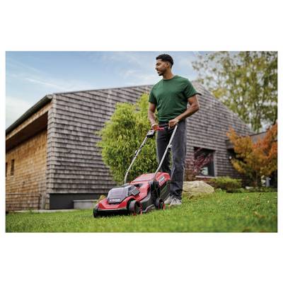 Un homme tondant la pelouse avec une tondeuse électrique rouge dans un jardin arrière, portant une chemise verte et un pantalon gris, avec une maison en bois et des arbres en arrière-plan.