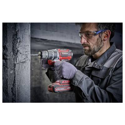 Une personne portant des lunettes de protection et des gants utilise une perceuse sans fil rouge et noire pour faire un trou dans un mur en béton.