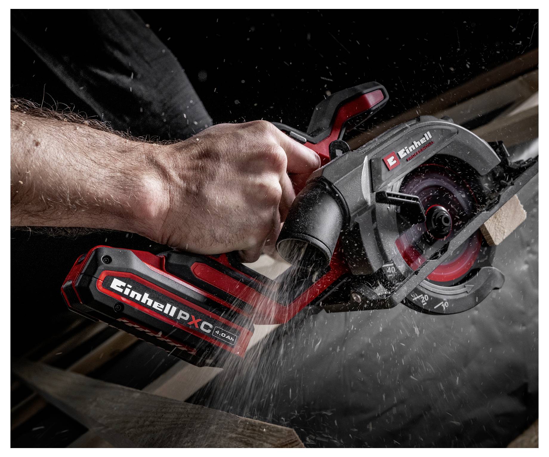 Une personne utilise une scie circulaire sans fil rouge et noire pour couper un morceau de bois, créant des copeaux de sciure visibles. La scie est étiquetée « Einhell PX-C ».