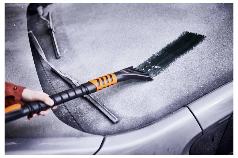 Une main utilise un grattoir à glace orange pour dégager le givre du pare-brise d'une voiture, révélant une bande nette par une matinée froide et givrée.