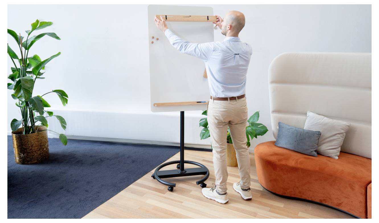 Une personne se tient dans un environnement de bureau moderne, écrivant sur un tableau à feuilles. Un canapé, des plantes et un tapis sombre sont présents.
