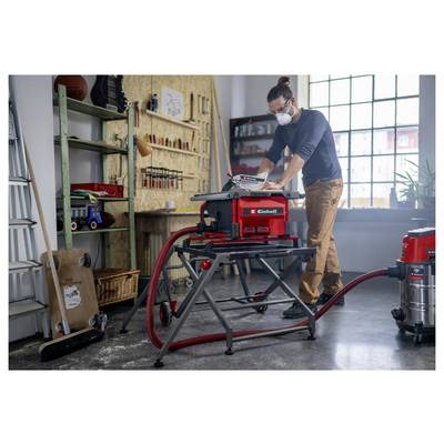 Une personne utilise une scie à table dans un atelier, portant un équipement de protection. Des outils et de l'équipement sont disposés autour, tandis que la lumière du soleil entre par une grande fenêtre.