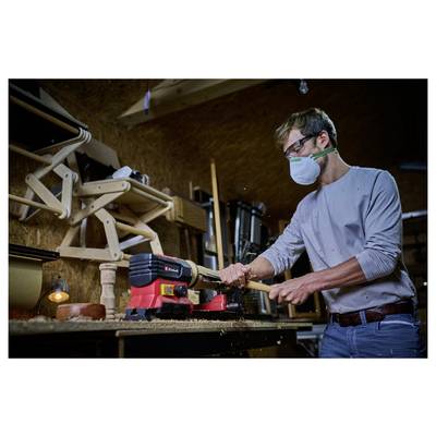 Une personne portant un masque utilise un tour à bois pour façonner du bois dans un atelier. Des outils et des structures en bois sont visibles en arrière-plan.