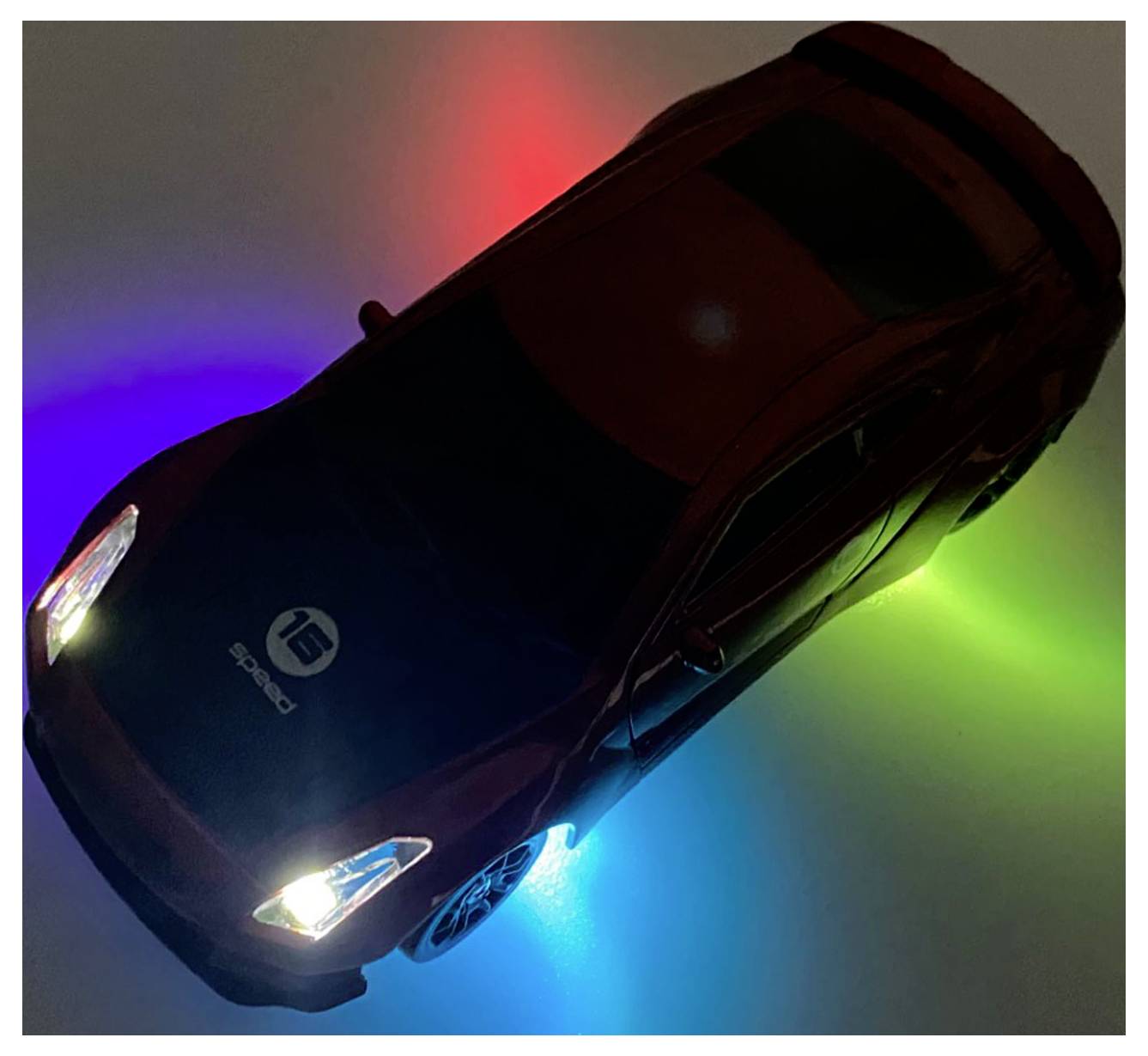 Une voiture miniature avec des phares lumineux et des lumières LED colorées en dessous, créant un effet d'auréole sur la surface en rouge, bleu et vert.