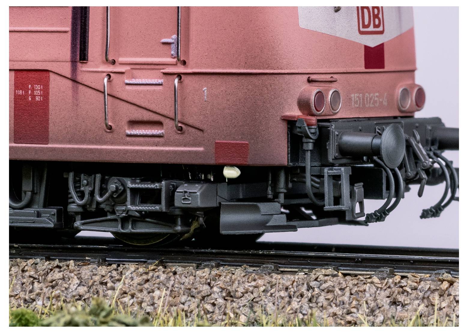 Gros plan sur l'arrière d'une locomotive rouge, montrant des pièces mécaniques détaillées et le logo 'DB'. Les rails sont visibles sous la locomotive.