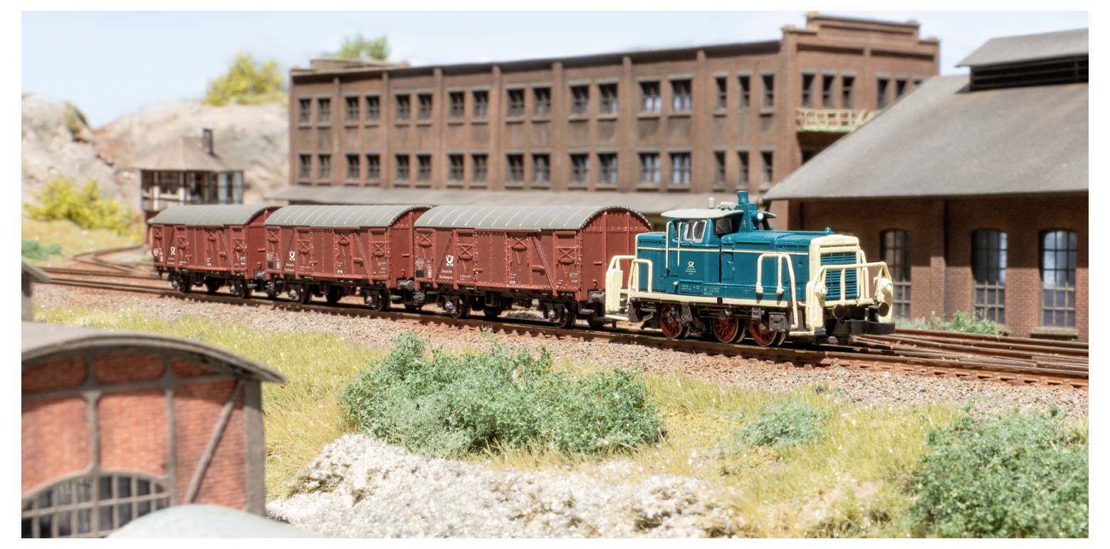 Un train miniature avec une locomotive bleue et des wagons de marchandises rouges passe devant un bâtiment industriel en briques et des paysages verdoyants dans un décor en miniature.