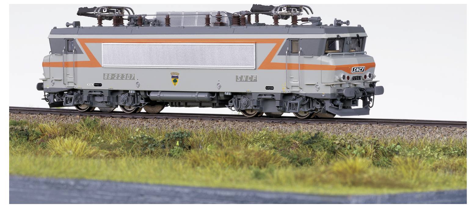Une locomotive de train miniature avec un design gris et orange exposée, circulant sur un rail entouré d'herbe et de paysage.