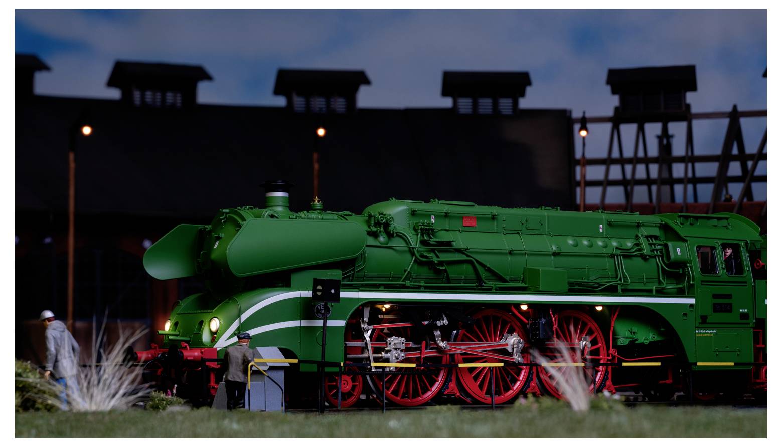 Un modèle de locomotive à vapeur verte avec des roues rouges est exposé dans un décor nocturne, avec un bâtiment et des personnages à proximité.