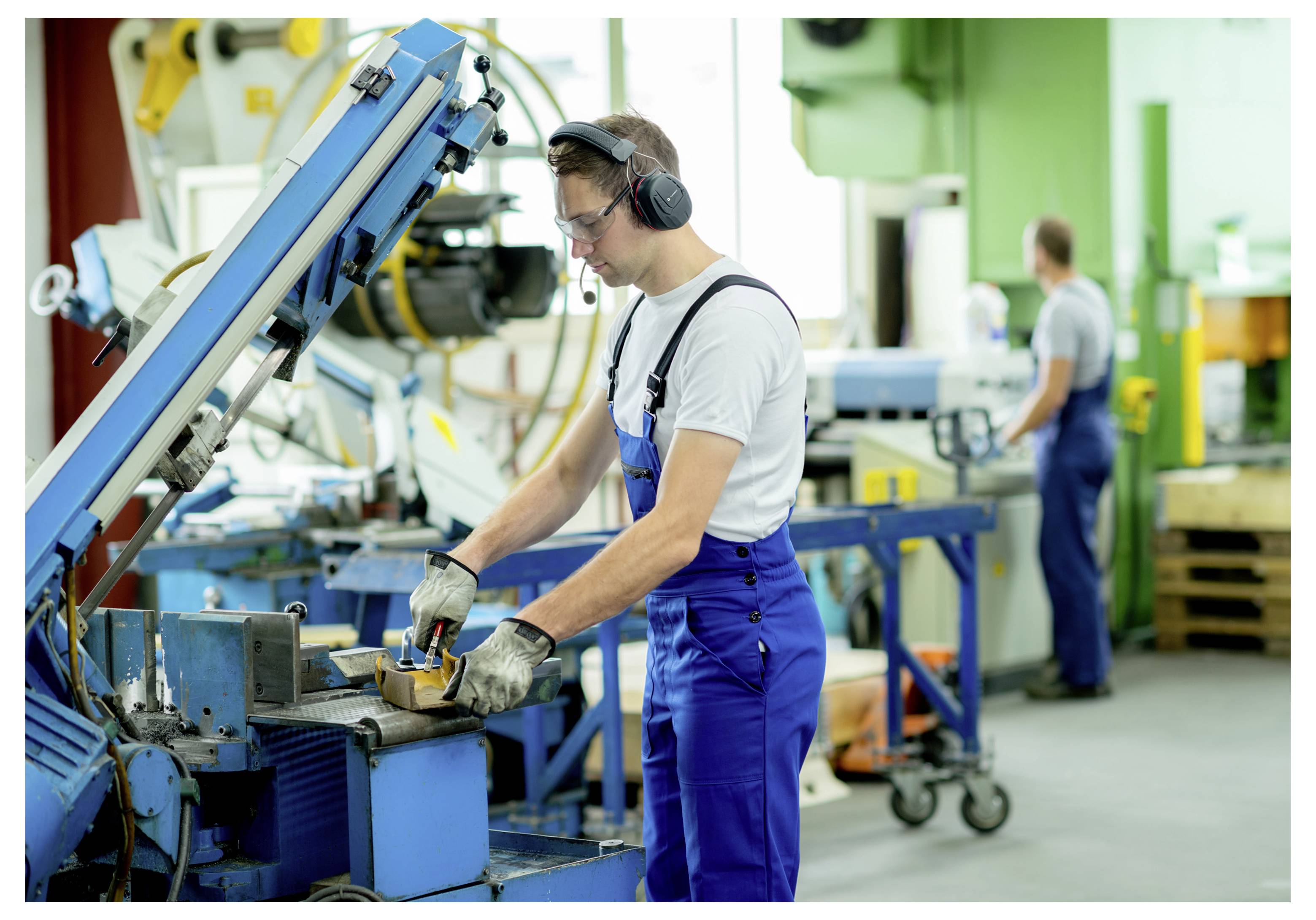 Ouvrier d'usine vêtu d'une salopette bleue et portant des écouteurs qui utilise une machine industrielle bleue dans un atelier, avec un autre ouvrier flou en arrière-plan.