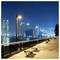 Un paysage urbain nocturne montrant une promenade en bord de mer avec des lampadaires, des bancs et une vue sur des gratte-ciels illuminés de l'autre côté de l'eau.