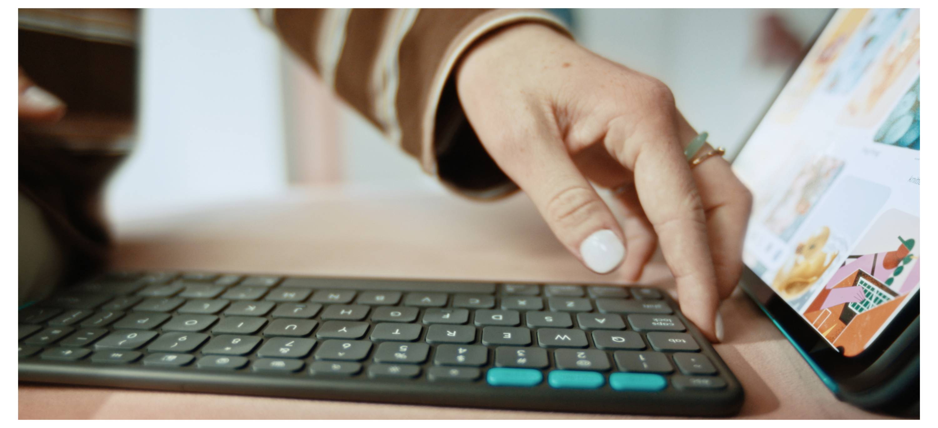 Une main d'une personne, portant un ongle peint en blanc, tend vers un clavier sans fil situé près d'une tablette affichant des images colorées.