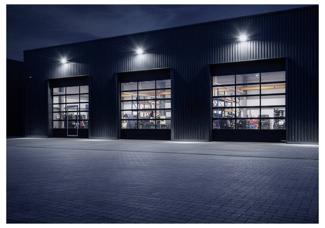 Un entrepôt bien éclairé à l'extérieur, de nuit, avec trois grandes portes de garage illuminées révélant des machines et de l'équipement à l'intérieur.