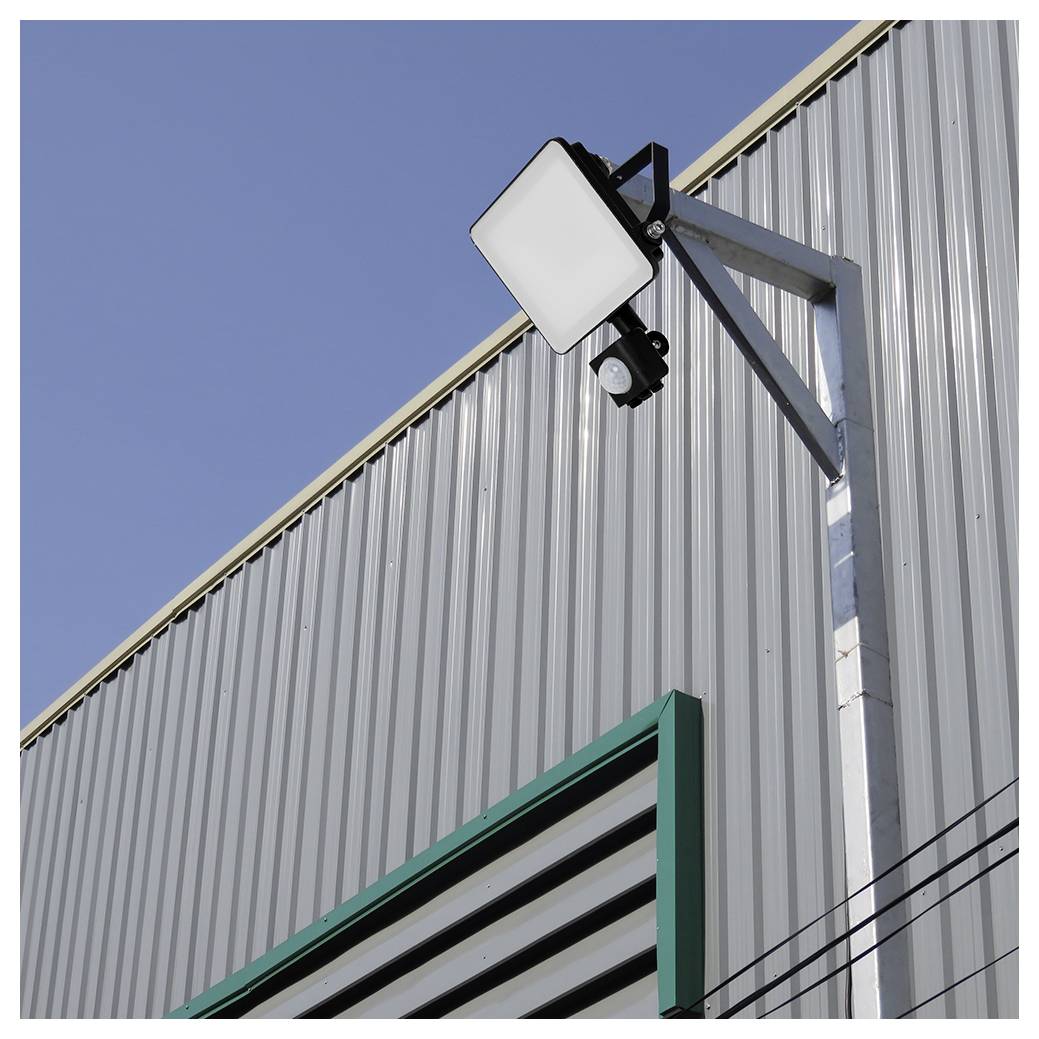 « Un projecteur de sécurité extérieur est installé sur un mur en tôle ondulée, positionné au-dessus d'une ouverture de ventilation au cadre vert, sous un ciel bleu dégagé. »