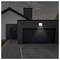 Une maison moderne avec un extérieur dont une seule lumière au-dessus de la porte du garage éclaire l'allée à la tombée du jour.