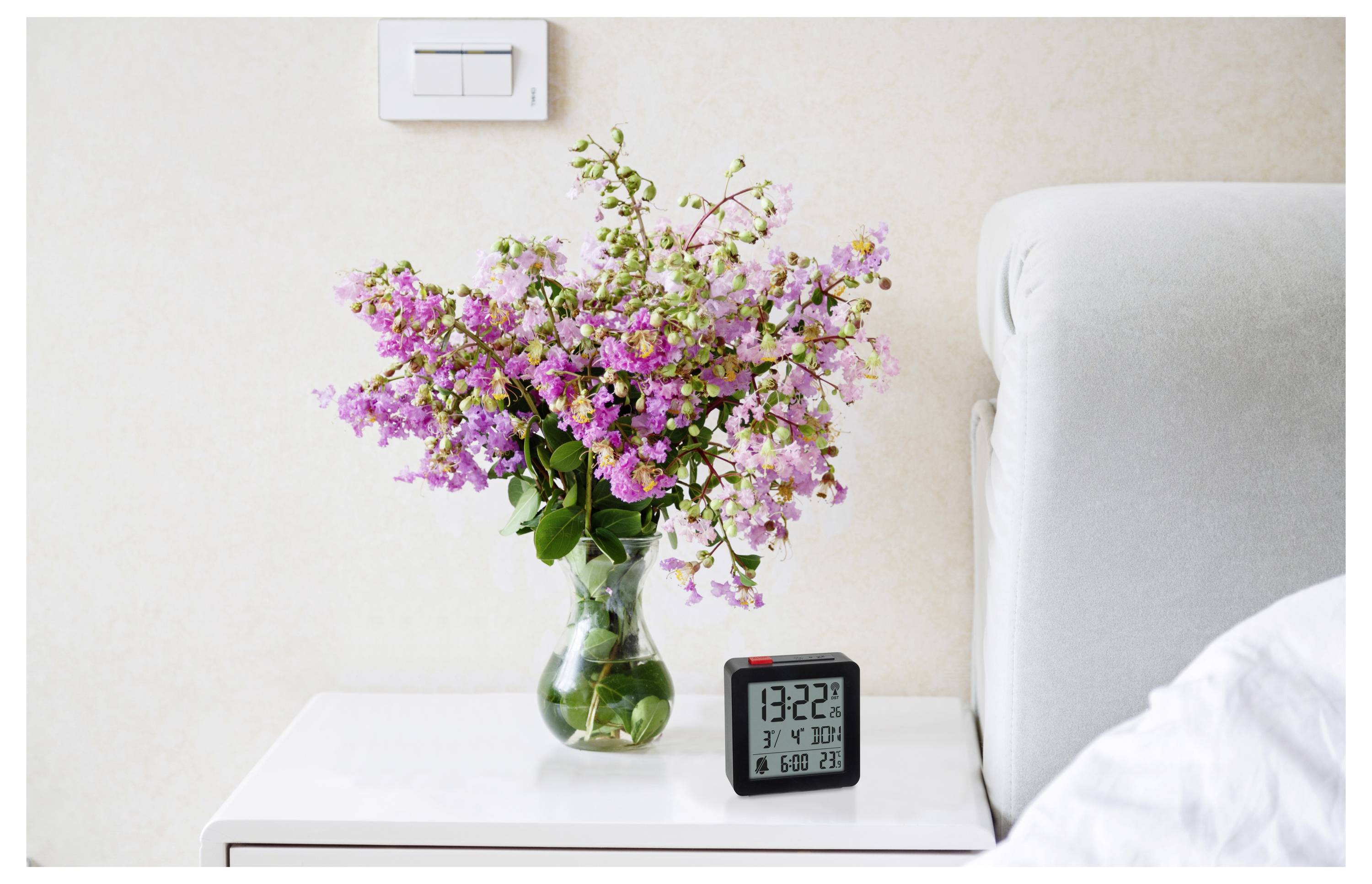 Un vase de fleurs roses sur une table de chevet à côté d'une horloge numérique affichant « 13:22 » et d'un interrupteur mural.