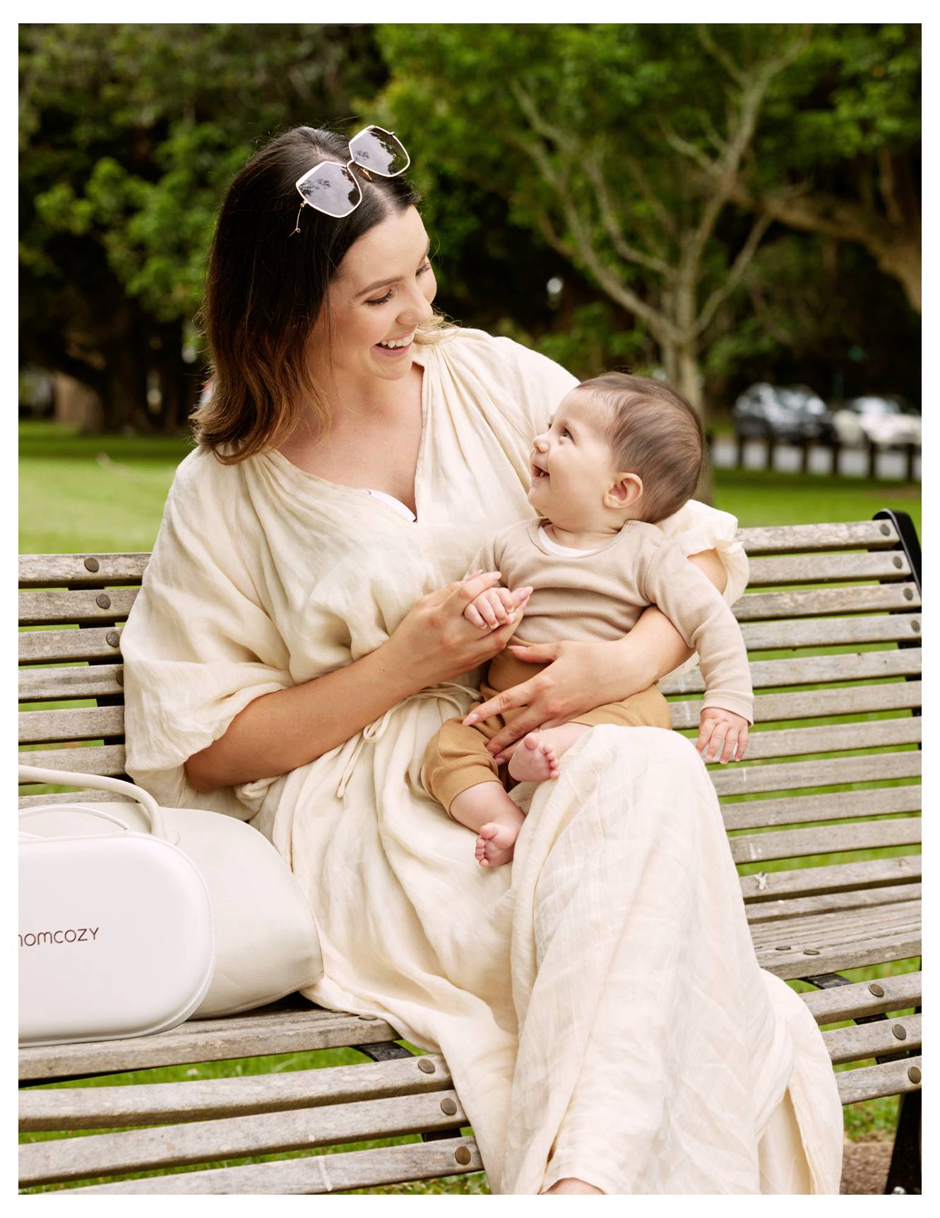 Une femme souriante en robe beige est assise sur un banc de parc, tenant un bébé riant habillé en beige, avec des arbres en arrière-plan.