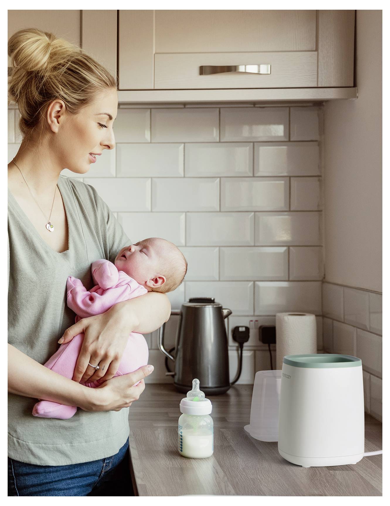 Une femme dans une cuisine tient un bébé habillé en rose. Un chauffe-biberon et un biberon sont posés sur le comptoir, suggérant qu'elle s'apprête peut-être à préparer un repas.
