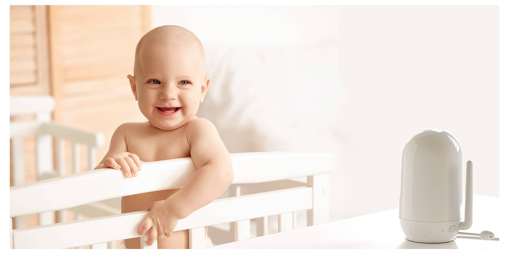 Un bébé souriant se tient debout dans son berceau, regardant un babyphone posé sur une table. La scène suggère un environnement sûr et joyeux.