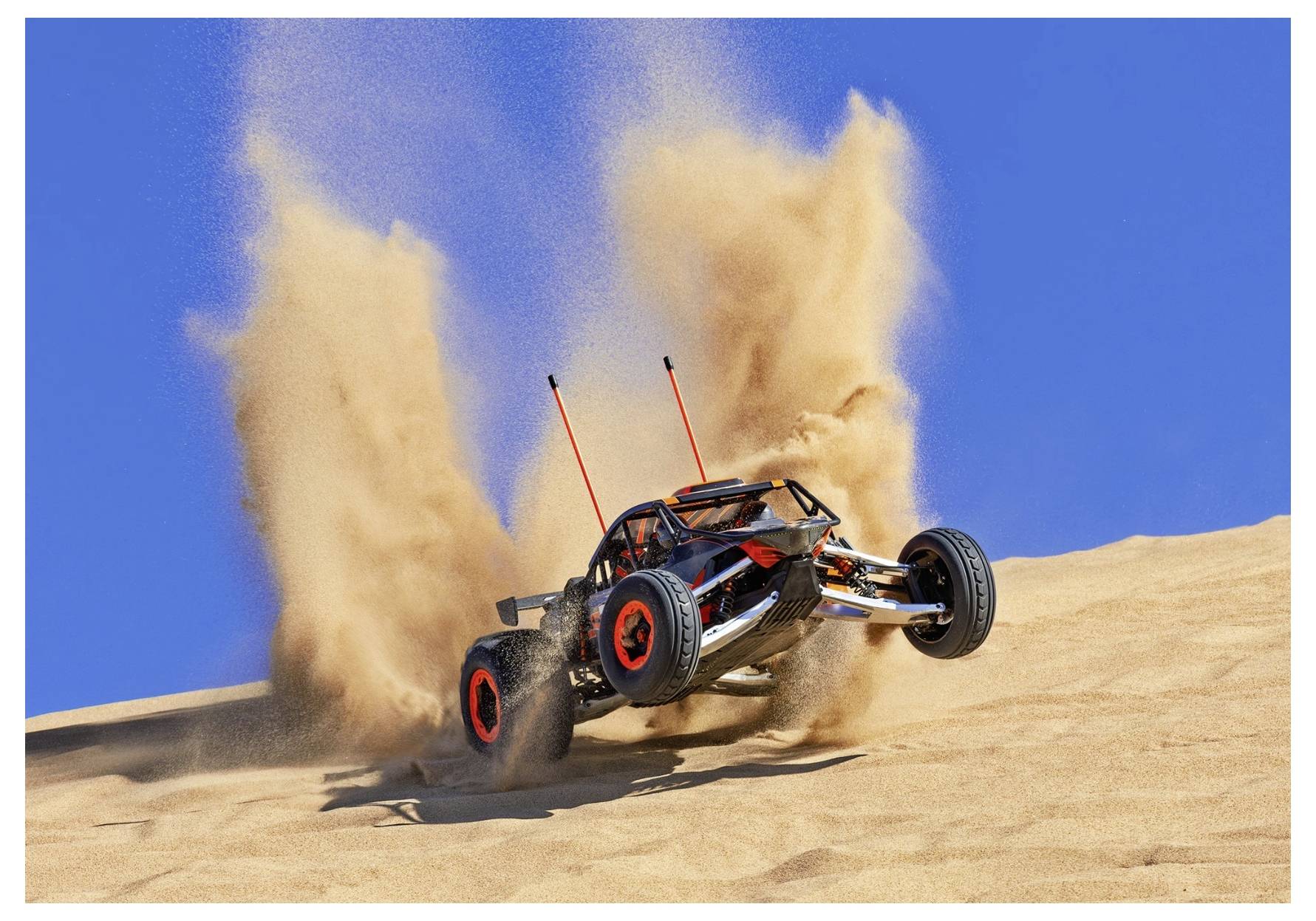 Une voiture télécommandée aux roues rouges soulève du sable en gravissant une dune désertique sous un ciel bleu dégagé.