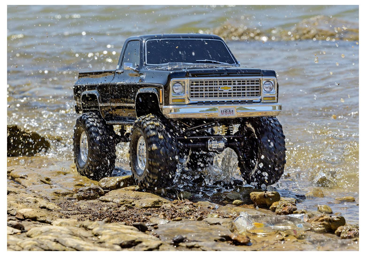 Un monster truck télécommandé roule sur un terrain rocailleux près d'un lac, projetant de l'eau autour de ses grandes roues.
