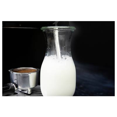 Une carafe en verre remplie de lait est en train d'être chauffée à la vapeur sur une machine à espresso, à côté d'un porte-filtre contenant du café, sur un fond sombre.
