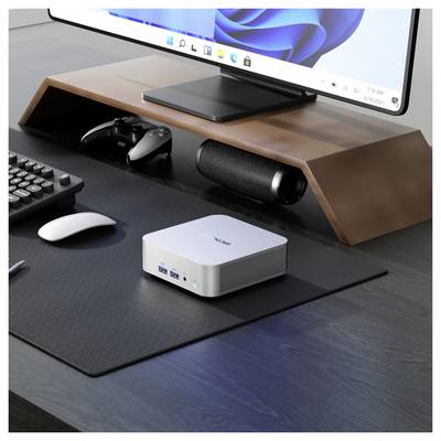 Un setup de bureau moderne avec un moniteur sur un support en bois, un clavier, une souris, des manettes de jeu, un mini ordinateur de bureau et un haut-parleur en dessous.