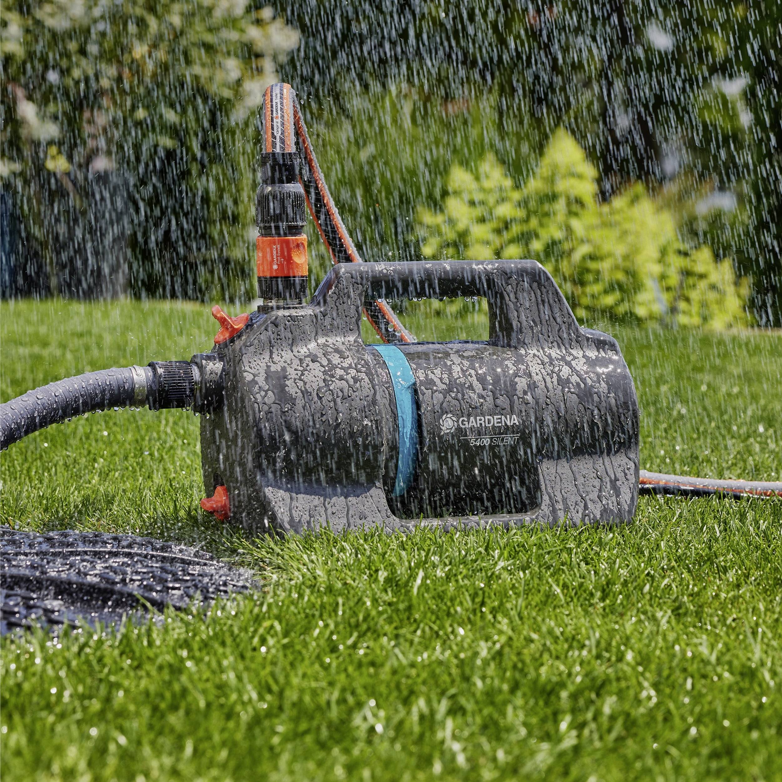 Une pompe à eau est sur une pelouse, projetant de l'eau sur l'herbe. La pompe est équipée d'un tuyau orange et noir, avec de la verdure en arrière-plan.