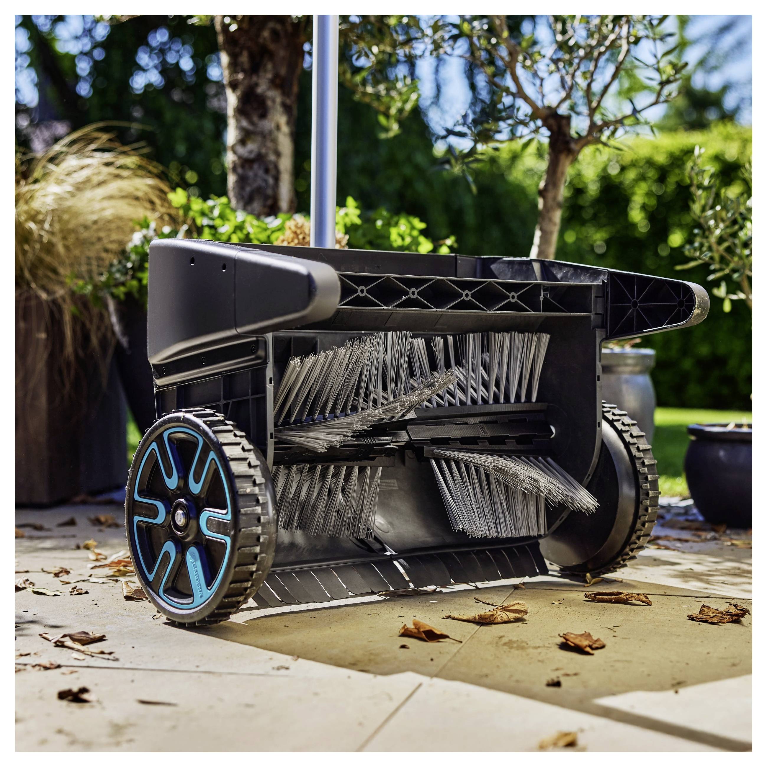 Un balai mécanique pour pelouse avec des brosses rotatives et un bac de collecte se tient sur un patio en pierre, entouré de plantes dans un décor de jardin.