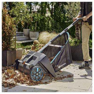 Une personne utilise un collecteur de feuilles de jardin sur une terrasse, rassemblant les feuilles tombées. L'outil est équipé de roues, avec un sac en maille pour la collecte des feuilles.